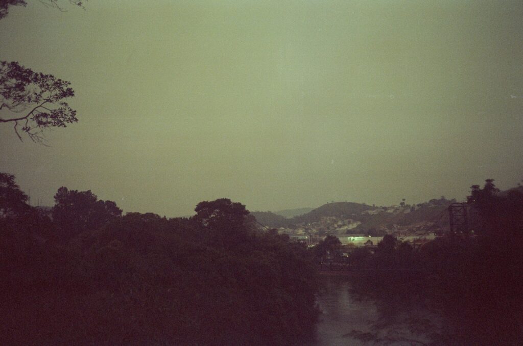 fotografia bem escura do rio Paraíba do Sul, captável apenas pelo brilho da água; matas densas ao lado, céu nublado, no horizonte, luzes e montanhas bem distantes.